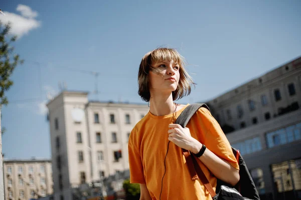 Jeune fille avec casque dans un t-shirt jaune et avec un sac à dos se promène le long d'une rue de la ville par une journée ensoleillée d'été — Photo