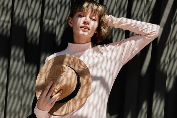 Menina elegante em um vestido suavemente rosa e chapéu de palha de pé com o chapéu de palha em sua mão ao lado de uma cerca de metal escuro na luz do sol com sombras refletidas — Fotografia de Stock