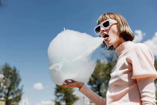Chica con estilo en un vestido suavemente rosa y gafas de sol blancas come un algodón de azúcar en la calle en un día soleado — Foto de Stock