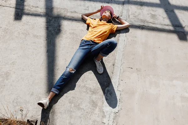Het meisje in geel t-shirt, jeans en roze vizier legt op een betonnen plaat op een zonnige dag buiten — Stockfoto