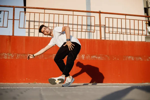 Kot giyinmiş ve beyaz t-shirt şık genç adam güneşli bir günde boyalı beton duvara karşı sokakta brakedance dans ediyor