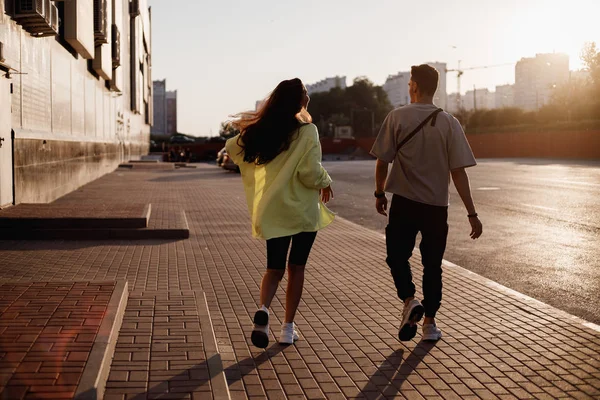 Pemuda bergaya pria dan gadis berjalan bersama-sama di alun-alun untuk parkir di samping bangunan pada matahari terbenam — Stok Foto
