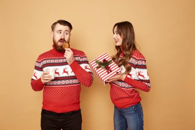 Noel hediyesi tutan kız ve elinde kurabiye ve kırmızı ve beyaz kazakgiymiş bir bardak süt tutan bir adam stüdyoda bej bir arka plan üzerinde geyik standı
