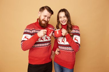 Gülümseyen kız ve geyik ile kırmızı ve beyaz kazak giymiş bir adam ellerinde kırmızı bardak tutuyor ve stüdyoda bej arka plan üzerinde durmak