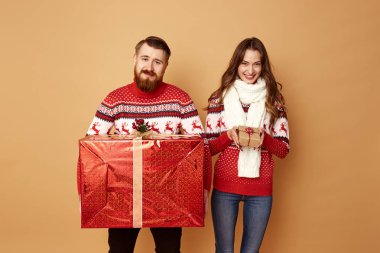 Kız sürpriz küçük bir hediye tutan ve adam stüdyoda bej arka plan üzerinde geyik standı ile kırmızı ve beyaz kazak giymiş büyük bir hediye tutan