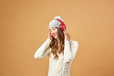 Kırmızı Kulaklığımda gülümseyen kız beyaz örgü Kazak giymiş ve şapka stüdyoda bej renkli bir arka plan üzerinde duruyor
