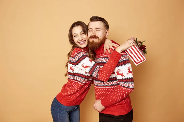 Дівчина задоволена хлопцем, який дарує їй різдвяний подарунок, обидва одягнені в червоні та білі светри з оленями і стоїть на бежевому тлі в студії — стокове фото