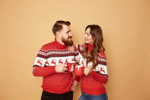 Smiling girl and a guy dressed in red and white sweaters with deer are holding red cups in their hands and stand on a beige background in the studio