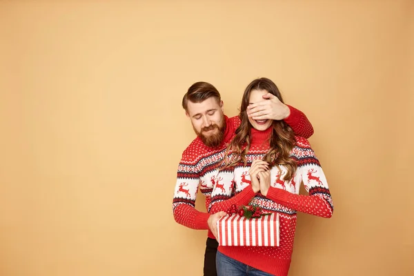 Ο τύπος κλείνει τα μάτια των κοριτσιών με το χέρι του και της κάνει δώρο ένα μπεζ φόντο στο στούντιο. Και οι δύο ντυμένοι με κόκκινα και άσπρα πουλόβερ με ελάφια. — Φωτογραφία Αρχείου