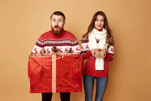 Το κορίτσι που κρατά ένα μικρό δώρο εκπλήσση και ο τύπος που κρατά ένα τεράστιο δώρο ντυμένος με κόκκινο και λευκό πουλόβερ με ελάφια σταθεί σε ένα μπεζ φόντο στο στούντιο — Φωτογραφία Αρχείου