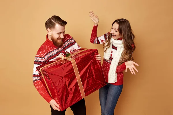 Дівчина дивно дивиться на хлопця, який тримає величезний подарунок. Вони обидва одягнені в червоні та білі светри з оленями — стокове фото