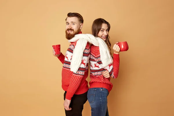 Дівчина і хлопець, одягнені в червоні та білі светри з оленями та білим в'язаним шарфом, стоять на спині і тримають червоні чашки на бежевому тлі — стокове фото