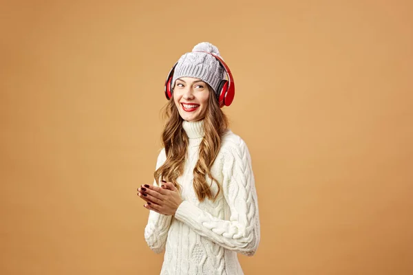 Menina sorridente em fones de ouvido vermelhos vestidos com suéter de malha branca e chapéu fica em um fundo bege no estúdio — Fotografia de Stock