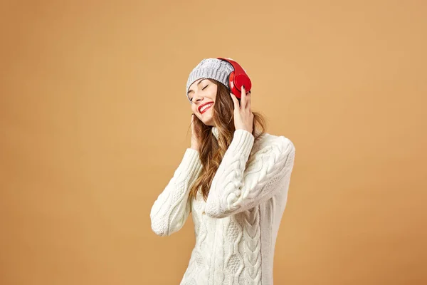 Lachende meisje in de rode hoofdtelefoon gekleed in witte gebreide trui en muts staat op een beige achtergrond in de studio — Stockfoto