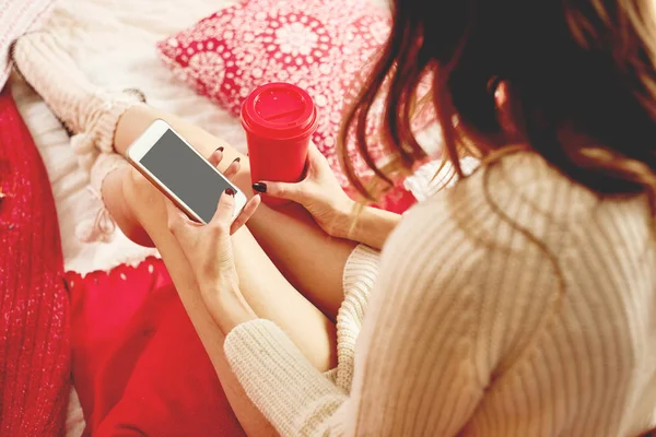 Girl dressed knitted dress and knitted socks lies on red-white blankets and pillows and holds a mobile phone and red cup of hot drink
