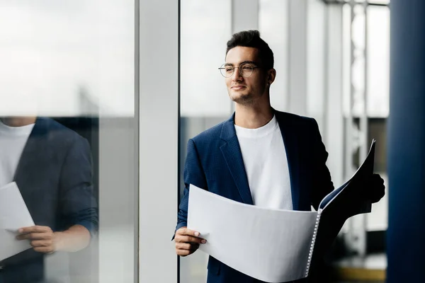 Arquitecto profesional vestido con ropa elegante está al lado de la ventana y mantiene la hoja con el dibujo en sus manos — Foto de Stock