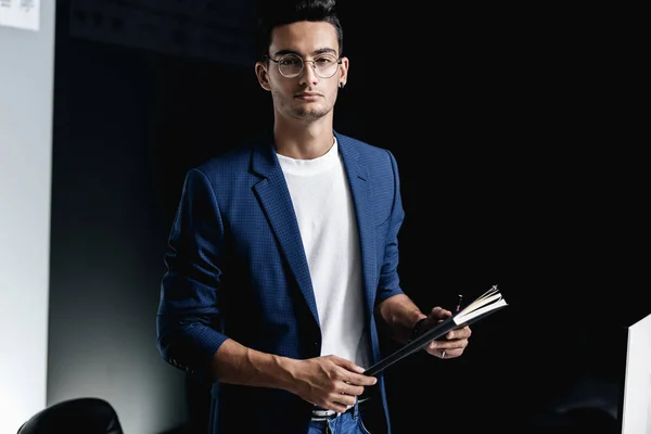 Élégant jeune architecte en lunettes vêtu d'une veste à carreaux bleu garde un carnet dans ses mains dans le bureau Image En Vente