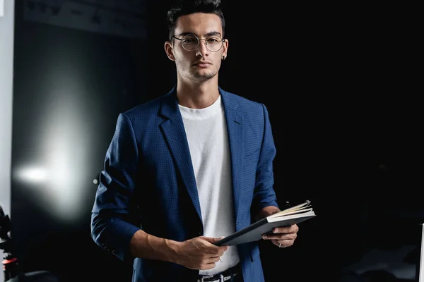 Stilvoller junger Architekt mit Brille im blau karierten Sakko hält im Büro ein Notizbuch in der Hand Stockfoto