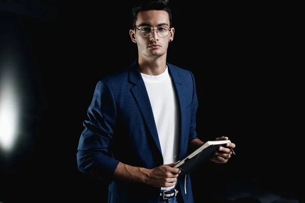 Stylowy młody architekt w okularach ubrany w niebieską kurtkę w kratkę trzyma notatnik w rękach w biurze Zdjęcia Stockowe bez tantiem