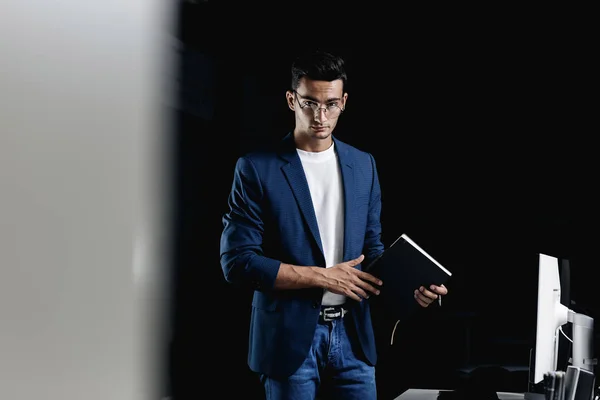 Arhitect tânăr elegant în ochelari îmbrăcați într-o jachetă albastră cu dame păstrează un carnețel în mâinile sale în birou Imagine de stoc