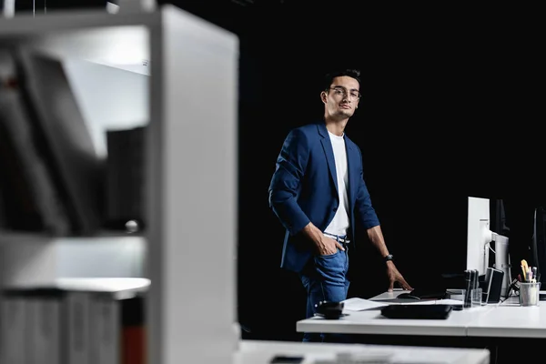 Architecte professionnel élégant en lunettes vêtu d'une veste à carreaux bleus se tient à côté du bureau avec ordinateur dans le bureau Photos De Stock Libres De Droits