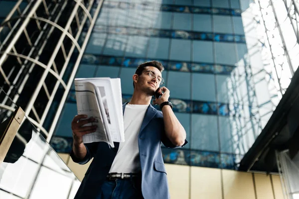Arhitect în haine elegante deține foaie cu desen în mână și vorbește telefonic pe fundalul unei clădiri moderne cu mai multe etaje din sticlă — Fotografie, imagine de stoc