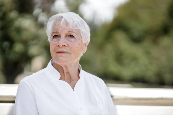 Élégante femme âgée aux cheveux gris dans la chemise blanche est assis sur le banc dans un parc par une journée chaude Photos De Stock Libres De Droits