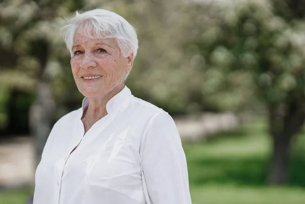 Uśmiechnięta, elegancka starsza kobieta w białej koszuli stoi w parku w ciepły słoneczny dzień Obrazy Stockowe bez tantiem