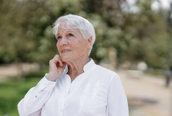 Elegante ältere grauhaarige Frau im weißen Hemd steht an einem warmen, sonnigen Tag in einem Park Stockbild