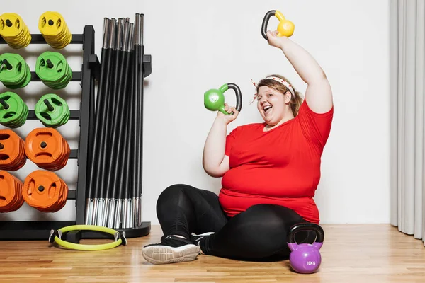 Dickes und lustiges Mädchen in Sportkleidung trainiert mit Hanteln in der Sporthalle neben anderen Sportgeräten Stockfoto