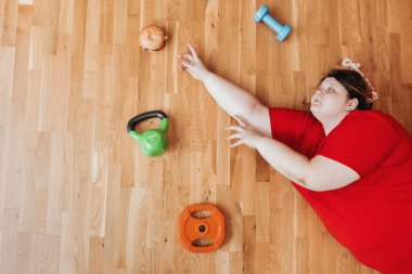 Spor kıyafetleri giymiş şişman ve komik bir kız kafasında bandajla spor malzemelerinin yanına uzanır ve ellerini burgerin üzerine koyar.