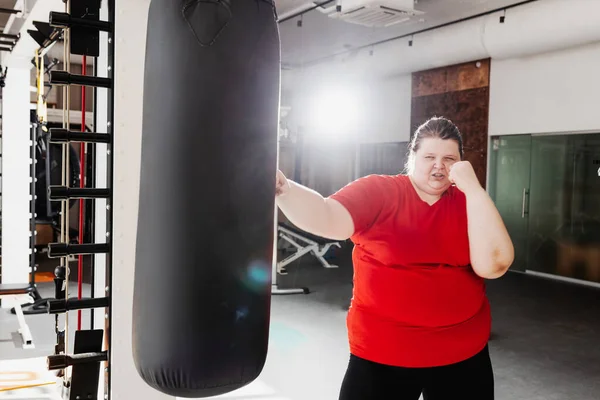 Fat girl in the sportswear is boxing punching bag in the gym