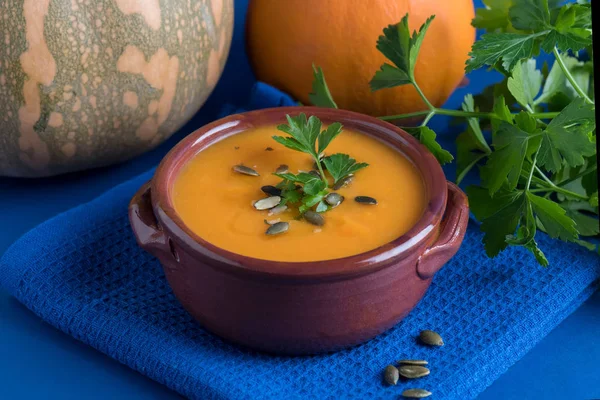 Веганский суп из тыквы в глиняной миске, подаваемый с петрушкой, оливковым маслом и тыквенными семечками на синем фоне — стоковое фото