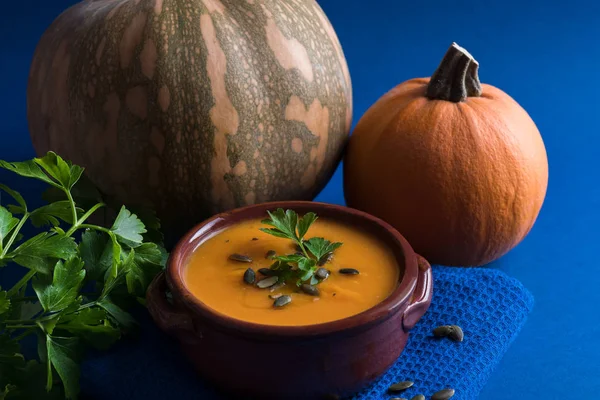 Close up of pumpkin soup in a bowl served with parsley, pumpkin seeds and two pumpkins on the blue bright background