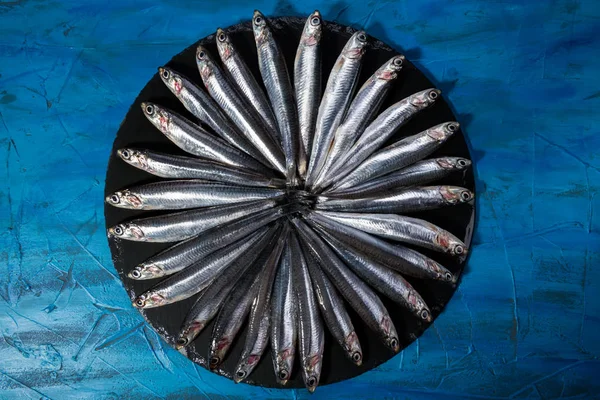Padrão de peixe. Anchovas na forma de um círculo em uma pedra preta em um fundo azul do mar. Frutos do mar. Pequenos peixes marinhos — Fotografia de Stock