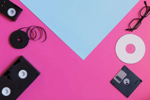 Retro storage devices: two videocassette, floppy disk, CD and glasses. Outdated technology concept on pink blue colored paper background, retro toned image, top view, minimalism