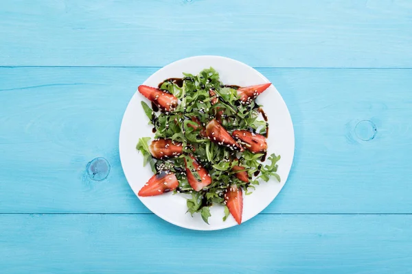 Salade diététique bio saine avec roquette, fraises et sésame avec glaçure balsamique dans un plateau blanc sur un fond en bois bleu pastel, vue de dessus — Photo