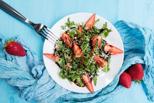 Здоровый органический диетический салат с рукколой, клубникой и кунжутом с бальзамической глазурью на белом пластине на пастельно-голубом фоне, вид сверху — стоковое фото