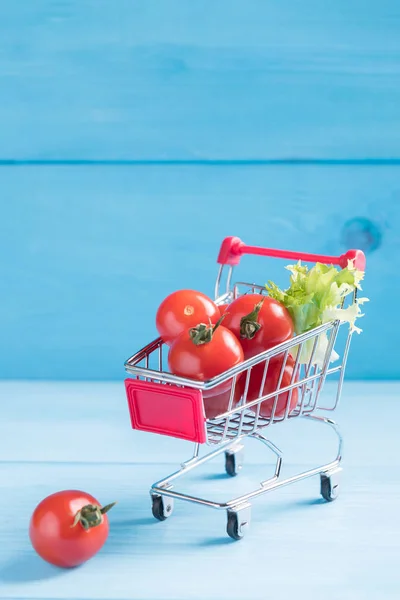 食品ミニマルコンセプト、コピースペース付きの青い木製の背景におもちゃのショッピングカートに自然な有機トマト — ストック写真