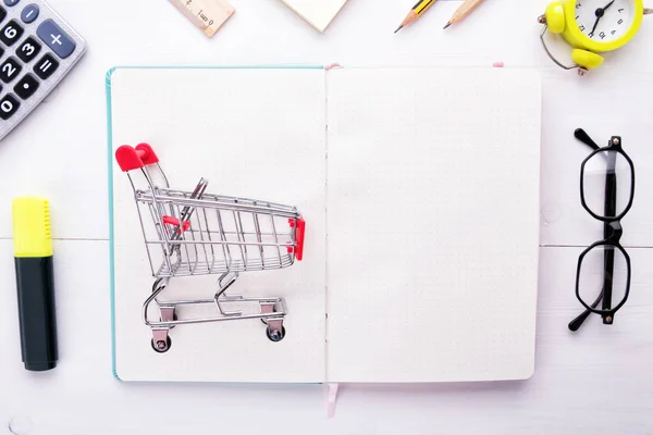 Organizar su lista de compras: pequeño carrito de comestibles en cuaderno limpio blanco o planificador con papelería sobre un fondo de madera blanco —  Fotos de Stock