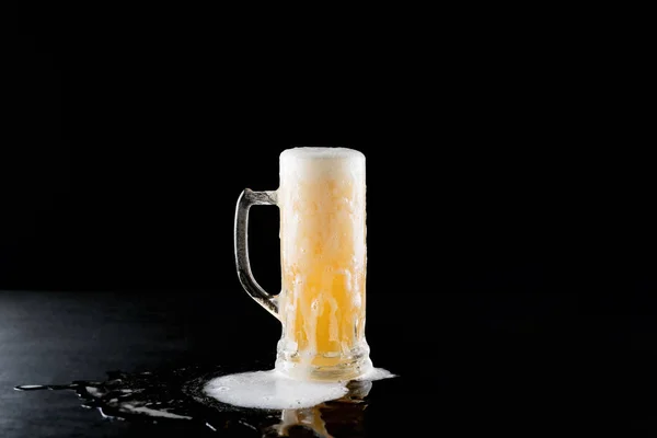 Overflowing mug of fresh cold light beer with froth flowing on the table and beautiful spilled beer on a black background with reflection — Stock Photo, Image