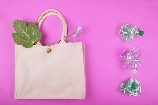 Reusable cotton bag with a glass bottle and a sheet of figs and three crumpled plastic bottles on a bright pink fuchsia background. Zero waste concept