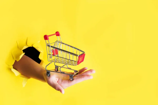 Mano femenina sostiene a través de un agujero un mini carrito de la compra de comestibles en un papel amarillo concepto de ventas de fondo con espacio de copia, minimalismo —  Fotos de Stock