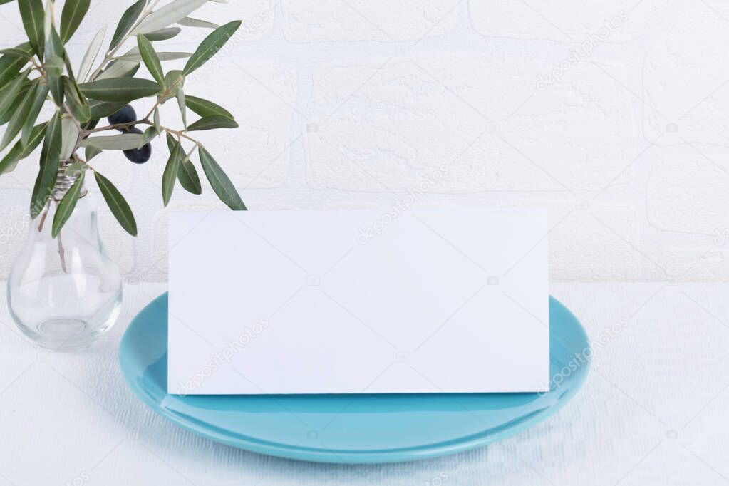 Minimalistic wedding composition with empty cards mock up for invitation, menu, place card on a pastel blue porcelain plate with olive branch on white background. top view stylish art concept