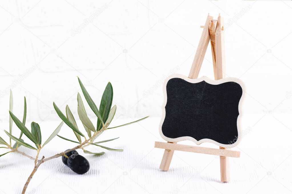 Blank outdoor chalk board stand mockup with olive branch on white brick wall background. Empty street signage or blackboard with wooden frame template