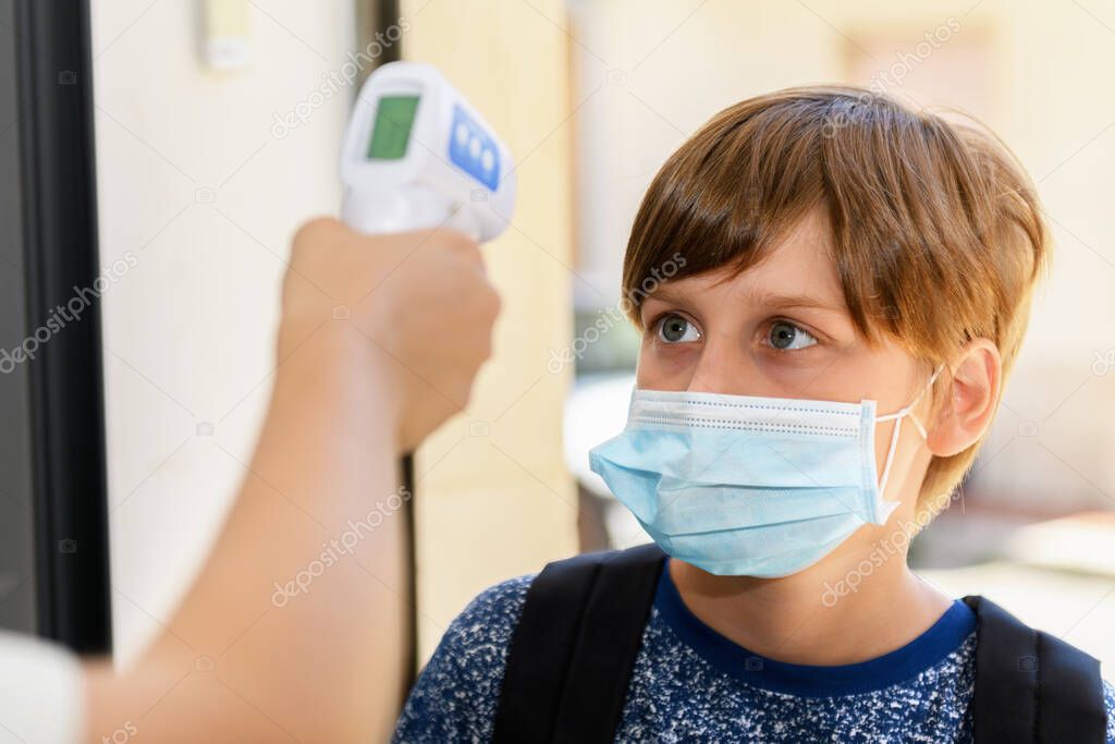 Teacher with facemask scanning a kid with digital thermometer, measuring body temperature before lessons. New normal, back to school during coronavirus pandemic.