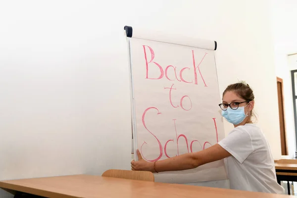 Young girl wearingmedical mask writing back to school on whiteboard in empty classroom, back to school during Covid-19 pandemic, new normal. Returning to school after quarantine, happiness and hope.