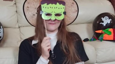 Gülümseyen bir kız kağıt maskeyle eğleniyor, komik ve korkunç suratlar takınıyor, Cadılar Bayramı için evde hazırlanıyor, Cadılar Bayramı 'nı evde şenlikli süslerle kutluyor.