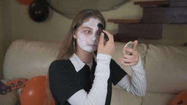 Bir kız Cadılar Bayramı için yüzünü beyaz makyajla boyuyor. Karnaval için ev makyajı, evde cadılar bayramı kutlaması.