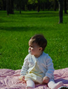 Parkta yeşil çim üzerinde pembe ekose parlak giysiler bebek. Yeni doğan bebek sıcak bir günde dışarıda. Küçük kız ilkbaharda yeşil bir çim enlaid üzerinde bir ekose üzerinde oynuyor